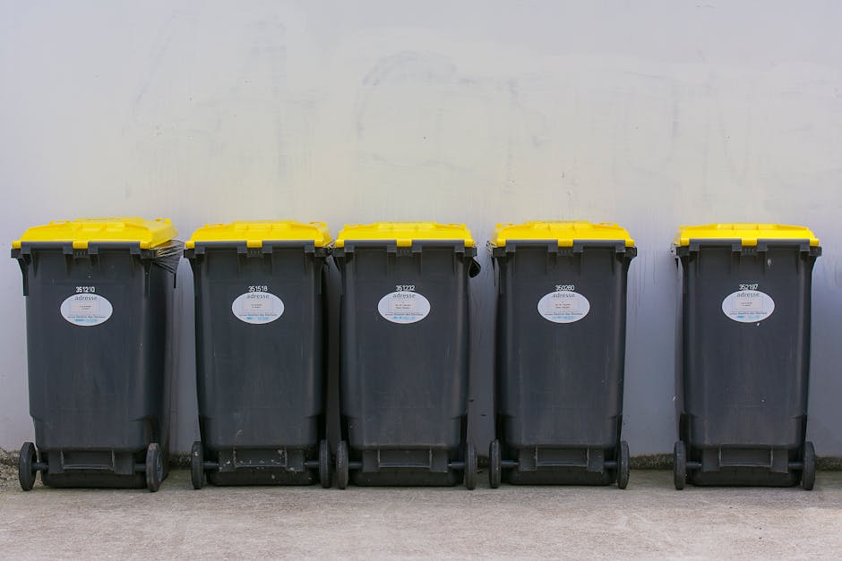 Close-up image of multiple black plastic rubbish bags tightly knotted at the top, stacked against a plain, light-colored wall. The bags are made from glossy polyethylene material, reflecting ambient l